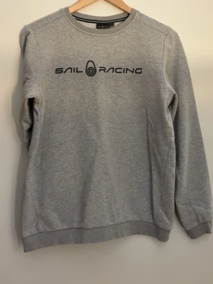 Grå sweatshirt från Sail Racing - Säljer en stilren grå sweatshirt från Sail Racing med svart logotyptryck på bröstet. Tröjan har rund halsringning, långa ärmar och ribbade muddar vid ärmslut och nederkant. Perfekt för en avslappnad look.