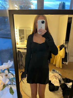 Svart långärmad kortklänning - Snygg svart klänning med lång ärm och fyrkantig urringning. Klänningen är kort och har en enkel, stilren design som passar perfekt till olika tillfällen. Materialet känns mjukt och följsamt mot kroppen.