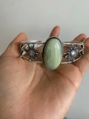 s925 Silvrigt armband med grön sten - Unikt armband i silverfärgad metall med en stor oval grön sten i mitten. På sidorna finns solformade detaljer med små pärlliknande inslag. Armbandet har en bohemisk och cool vibe, perfekt för att lyfta din stil.main material: sterling silver pläterat s925 justerbart 