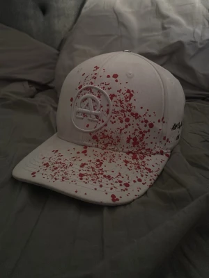 Vit keps med röd färgstänk All Caps One - Säljer en vit keps från All Caps One med unikt rött färgstänksmönster över hela framsidan och skärmen. Kepsen har broderad logga framtill och text på sidan. Justerbar baksida och klassisk form. Perfekt för dig som vill sticka ut med din stil.