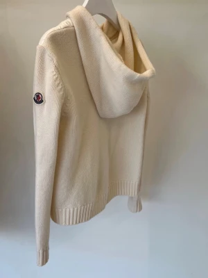 Moncler cardigan  - Säljer en beige hoodie från Moncler med stickade ärmar och quiltad framsida. Tröjan har dragkedja, huva och Moncler-logga på ärmen. Perfekt för dig som gillar exklusiv stil. Materialmix med både stickat och vadderat tyg. Bra skick, men har nån defekt skriv för fler bilder eller frågor💕