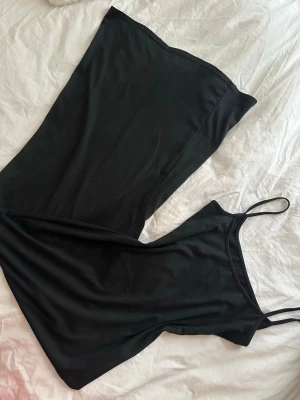 Svart lång slipklänning - Säljer en svart lång slipklänning med tunna axelband och rak passform. Klänningen har en minimalistisk look och är gjord i ett mjukt, stretchigt material som känns skönt mot huden. Perfekt för dig som gillar enkel och stilren design.