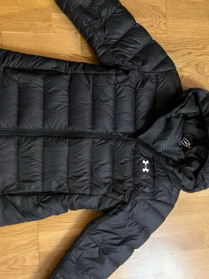 Svart dunjacka från Under Armour - Snygg svart dunjacka från Under Armour med huva och dragkedja. Jackan har ett diskret mönstrat innerfoder och det klassiska Under Armour-loggan på bröstet. Perfekt för kalla vinterdagar och riktigt skön att bära.