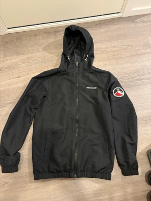 Svart vindjacka från Ellesse - Svart vindjacka från Ellesse med huva och dragsko, hel dragkedja framtill och två sidofickor. Jackan har resår vid ärmslut och midja samt en snygg patch på ena ärmen. Perfekt för blåsiga dagar och streetwear-stil.