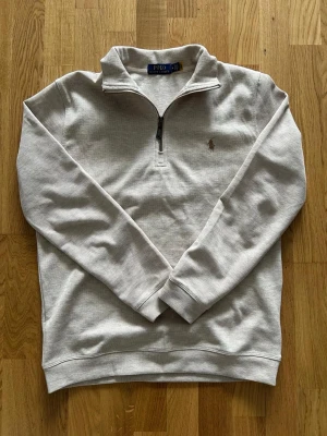 quarter zip från Polo Ralph Lauren - Snygg tröja med quarter zip och hög krage från Polo Ralph Lauren. Tröjan har det klassiska broderade logotypen på bröstet och ribbade muddar vid ärmslut och nederkant. Perfekt för en clean och stilren look.
