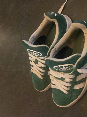 Gröna Adidas sneakers i mocka, adidas campus - Gröna Adidas campus i mocka med vita detaljer och klassiska tre ränder på sidan. Skorna har vita snören och Adidas logga på plösen. Låg modell som passar perfekt till streetwear och vardagsstil.