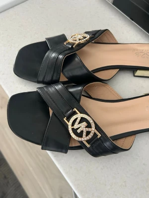 Svarta sandaler från Michael Kors - Stilrena svarta sandaler från Michael Kors med breda remmar i läder, fyrkantig tå och guldfärgade detaljer. Dekorerade med MK-logga i guld och strass framtill samt guldfärgad kant på den låga klacken. Perfekta för en trendig look.