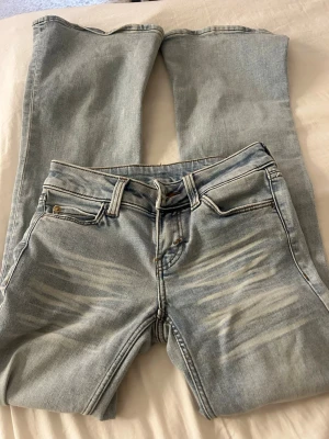 Ljusblå jeans från Cheap Monday - Säljer ett par ljusblå jeans från Cheap Monday med coola slitningar framtill och en grön patch med logga bak. Klassisk femficksmodell och raka ben. Perfekta för dig som gillar streetwear och vill ha en avslappnad look.