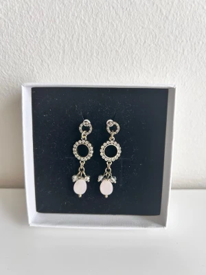 Hängande örhängen med rosa pärlor - Säljer ett par hängande örhängen i silverfärg med cirkelformade detaljer och små gnistrande stenar. Örhängena har ljusrosa pärlor och små kristalliknande dekorationer som ger en elegant look. Perfekt för dig som gillar unika accessoarer.