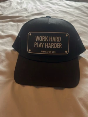 Svart truckerkeps John Hatter & Co - Snygg svart truckerkeps från John Hatter & Co med mesh på sidorna och böjd skärm. Framtill sitter en skylt med texten 'WORK HARD PLAY HARDER' i vitt. Kepsen har justerbar snapback och cool streetwear-vibe. Den är som ny då jag köpte den men ångra mig typ direkt så har ej använt den