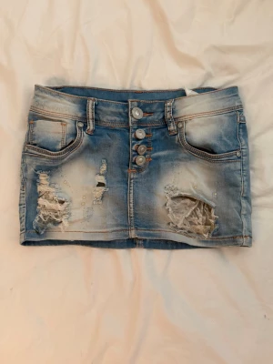Lågmidjad jeanskjol - Säljer en kort, ljusblå jeanskjol med slitna detaljer och fransar framtill. Köpte den på en marknad i Tyskland för ca ett år sedan men har aldrig använt den själv då den är lite för liten för mig! 💕