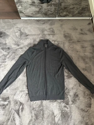 Mörkgrå ziptröja med hög krage - Snygg mörkgrå ziptröja med hel dragkedja och hög krage. Tröjan har en minimalistisk design och är tillverkad i ett mjukt, tunt material som passar perfekt för lager-på-lager. Långa ärmar och ribbade muddar ger en clean look.