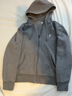 Grå hoodie från Polo Ralph Lauren - Snygg grå hoodie från Polo Ralph Lauren med dragkedja, huva med dragsko och klassisk vit logga på bröstet. Tillverkad i mjukt bomullsmaterial och har två stora fickor framtill. Perfekt för en chill och avslappnad stil.