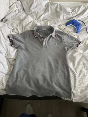Grå pikétröja från Moncler - Snygg grå pikétröja från Moncler med klassisk krage och knappar framtill. Tröjan har vita och röda detaljer på krage och ärmslut samt Moncler-logga på bröstet. Perfekt till en avslappnad men stilren look.