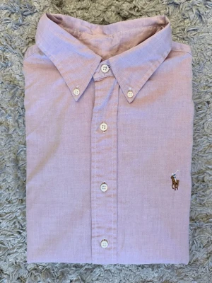 Ljusrosa skjorta från Ralph Lauren - Klassisk ljusrosa skjorta från Ralph Lauren med button-down krage och broderad logga på bröstet. Skjortan har lång ärm, vita knappar och är tillverkad i mjuk bomull. Perfekt för en clean och stilren look.