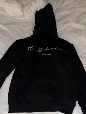  Han Kjøbenhavn - Säljer en svart hoodie från Han Kjøbenhavn med broderad text på bröstet och klassisk känguruficka. Hoodien har dragsko i huvan och ribbade muddar vid ärmslut och nederkant. Perfekt för en avslappnad och stilren look.