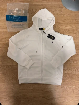 Vit hoodie från Polo Ralph Lauren - Säljer en fräsch vit hoodie från Polo Ralph Lauren med dragkedja, huva och snören. Klassisk logga broderad på bröstet och två fickor framtill. Perfekt för dig som gillar stilrena och tidlösa plagg. Materialet känns mjukt och skönt mot huden.