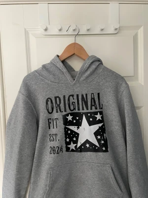 Grå hoodie med stjärntryck - Säljer en grå hoodie med svart och vitt tryck framtill där det står 'ORIGINAL FIT EST. 2024' och en stor stjärna med mindre stjärnor runt om. Hoodien har huva och magficka. Perfekt för en chill och avslappnad stil. Från SCNDI. Storlek S.
