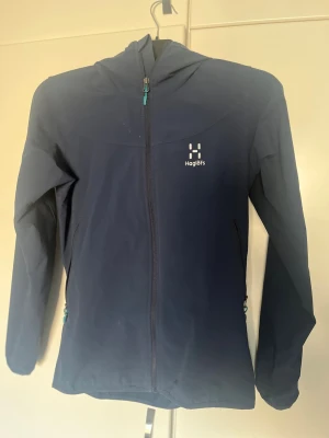 Mörkblå vindjacka från Haglöfs - Snygg mörkblå vindjacka från Haglöfs med hel dragkedja framtill och huva. Jackan har två sidofickor med dragkedja och diskret logga på bröstet. Perfekt för blåsiga dagar och outdoor-äventyr. Lätt och smidig design i slitstarkt material.