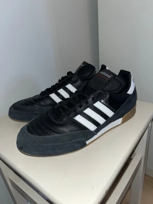 Adidas Mundial Team svart/vit sneaker - Säljer ett par Adidas Mundial Team sneakers i svart läder med vita klassiska stripes och detaljer. Skorna har en låg profil, svart mocka runt tån och gummisula. Perfekta för dig som gillar retrostil och sportig vibe. Använt de till futsall ett par gånger bara.