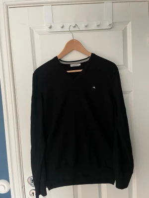 Svart stickad tröja från J.Lindeberg 100% merinoull - Klassisk svart stickad tröja från J.Lindeberg med V-ringning och diskret logga på bröstet. Tröjan har långa ärmar och ribbade muddar. Perfekt basplagg i garderoben, enkel att matcha till olika stilar. Tröjan är i sltorleken L men passar M. 100% merinoull