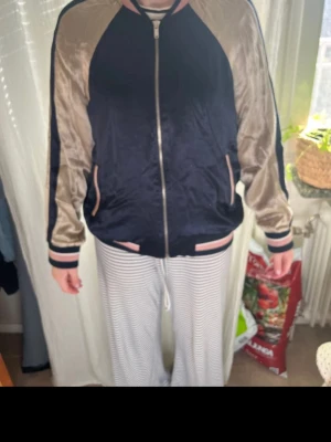 Svart och beige bomberjacka med brodyr - Snygg bomberjacka i svart och beige med glansigt material och blommig brodyr på ryggen. Fint skick!