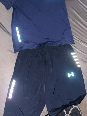 Svarta träningsshorts Under Armour - Snygga svarta träningsshorts från Under Armour med reflexdetaljer och logga på ena benet. Shortsen har snörning i midjan och är tillverkade i ett lätt och snabbtorkande syntetmaterial. Perfekta för gymmet eller löprundan.T-shirten är XL fast passar som L och shortsen är 2xl fast passar som xl 