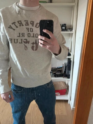 Beige sweatshirt från Polo RL Club - Säljer en beige sweatshirt från Polo RL Club med tryck i brunt framtill. Tröjan har rund halsringning, ribbade muddar och en liten patch nertill. Tillverkad i mjukt bomullsmaterial och har en avslappnad passform. Perfekt för en chill och stilren look.
