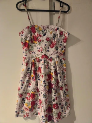 Blommig sommarklänning med smala axelband - Superfin kort klänning med blommigt mönster i rött, gult, blått och grönt på vit botten. Klänningen har smala axelband och en lätt utställd kjol, perfekt för varma dagar. Tillverkad i mjuk bomull som känns skön mot huden.