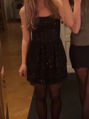 Svart glittrig kort klänning - Supersnygg svart kort klänning med glittriga detaljer. Klänningen är ärmlös och har en figurnära överdel med utställd kjol. Perfekt för dig som vill sticka ut och glänsa på nästa event!