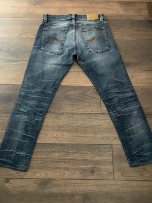 Blå slitna jeans från Nudie Jeans - Säljer ett par blå jeans från Nudie Jeans med slitningar och hål på framsidan. Jeansen har raka ben, klassiska bakfickor med Nudie-söm och är tillverkade i denim. Perfekta för en avslappnad och trendig look.