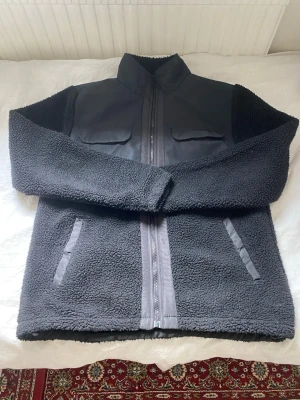 Svart teddyjacka från Saligia - Mysig svart teddyjacka från Saligia i storlek M. Jackan har hög krage, två bröstfickor med lock och två sidofickor. Materialet är fluffigt teddytyg med detaljer i slät textil på bröstet och runt dragkedjan. Perfekt för kalla dagar och riktigt snygg till streetwear. Pris kan diskuteras 