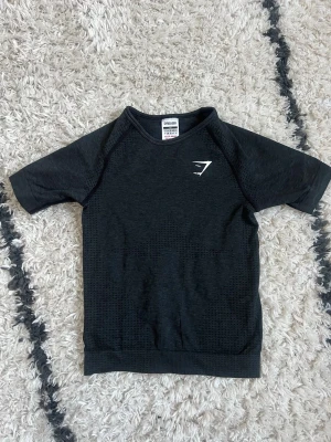 Gymshark 🖤 - Snygg mörkgrå / svart träningströja från Gymshark med korta ärmar och diskret mönstrad struktur. Tröjan har en vit Gymshark-logga på bröstet och är tillverkad i ett lätt, stretchigt funktionsmaterial som andas. Perfekt för gymmet eller löprundan. 