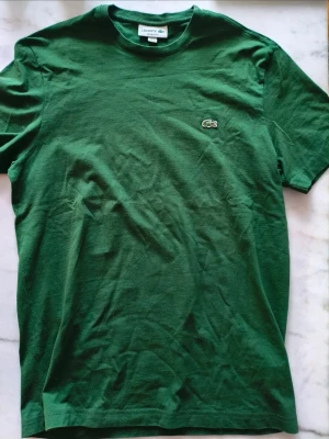 Grön Lacoste t-shirt med logga - Säljer en klassisk grön t-shirt från Lacoste med den ikoniska krokodilloggan broderad på bröstet. T-shirten har rund hals och korta ärmar, perfekt för en avslappnad och stilren look. Materialet är mjuk bomull som känns skönt mot huden.Tshirten är som ny.
