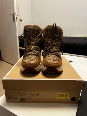 Bruna kängor från Michael Kors - Snygga bruna kängor från Michael Kors med MK-logga över hela ovandelen. Kängorna har en chunky vit sula, bruna mockadetaljer och snörning framtill. Perfekta för dig som vill ha både stil och komfort i höst och vinter Använts 1 gång för att prova om dom passade bra