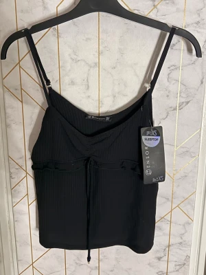 Svart ribbad linnetopp från New Yorker - Snygg svart linnetopp från New Yorker med tunna justerbara axelband och ribbad struktur. Toppen har en liten knytning framtill och är croppad i modellen. Köpt för 95kr.