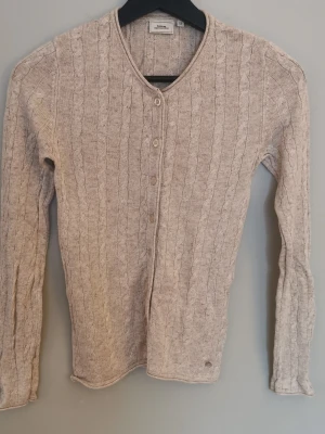 Melerad beige kabelstickad kofta från Boomerang  - Snygg beige kabelstickad kofta från Boomerang med rund hals och knappar framtill. Koftan har långa ärmar och passar perfekt till ett par jeans. Klassisk design med diskret logotyp vid nederkanten (har lossnat lite). Är i fint använt skick. Storlek XS