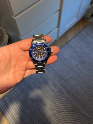 Blå automatisk klocka från Martyn - Snygg automatisk klocka från Martyn med blå urtavla och silverfärgat armband i metall. Urtavlan har ett coolt skelettmönster som visar mekaniken inuti och tydliga markeringar runt hela boetten. Perfekt för dig som gillar stilrena accessoarer.