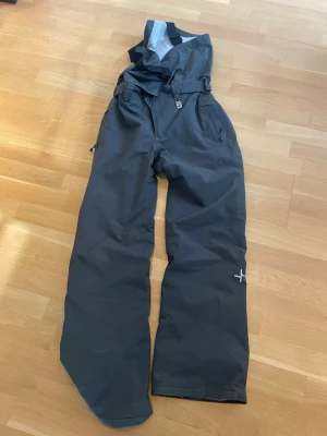 Cross bib skidbyxa - Säljer ett par svarta täckbyxor med hängslen och justerbar midja. Byxorna har dragkedjor vid fickorna och en liten vit detalj på ena benet. Perfekta för vinterns alla äventyr i snön.