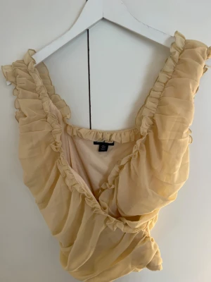 Beige omlottopp med volanger - Supersöt beige omlottopp från Primark med volangdetaljer längs kanterna. Toppen är ärmlös och har en luftig, romantisk look som passar perfekt till sommaren. Tillverkad i ett tunt, syntetiskt material som ger ett skirt intryck.
