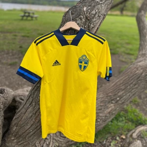 Sverige Hemmatröja 2020 #11 Guidetti - Gul kortärmad fotbollströja från Adidas med blå detaljer, officiella svenska landslagsemblemet och tryck 'Guidetti 11' på ryggen. Klassisk V-ringning och blå ränder på axlar och ärmslut. Tillverkad i lätt och ventilerande material. Defekt på sidan.
