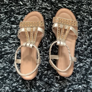 Glittriga sandaler i guld med remmar - Snygga sandaler i guld med glittrande stenar på remmarna och flätad detalj runt vristen. Platt sula och öppen tå, perfekta för sommaren när du vill sticka ut lite extra. Skorna har en elegant och trendig look med metallic-finish.