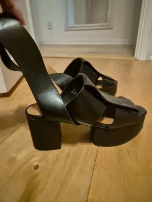 Svarta platåsandaler med bred klack st 36 - Säljer ett par svarta sandaler med breda korslagda remmar och hög platåsula. Skorna har chunky klack och öppen tå, vilket ger en cool och trendig look. Tillverkade i äkta läder och i fint skick!