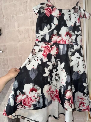 Blommig off-shoulder klänning  - Superfin blommig klänning med bar axel och smala axelband. Klänningen är något längre baktill och kortare framtill, vilket ger ett fantastiskt fall.  Detaljer: Off-shoulder med axelband, figursydd midja, utställd kjol. Bredd (axel till axel): 54 cm Längd nederdel (midja till fåll): 53 cm fram / 61 cm bak