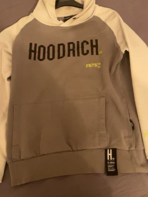 Grå och vit hoodie från Hoodrich - Säljer en snygg hoodie från Hoodrich i grått och vitt med stor magficka och svart HOODRICH-tryck på bröstet. Har huva och ribbade muddar. Detaljer i gult med FNTS-tryck och etikett nertill. Perfekt för en streetwear-look.