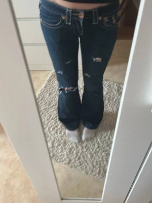 Mörkblå bootcut jeans med slitningar - Säljer ett par mörkblå bootcut jeans från True Religion med vita kontrastsömmar och slitna detaljer på benen. Jag är 161 och dom sitter ganska bra. Jeansen har låg midja, fickor med lock och knappar bak samt klassisk True Religion-lapp. Perfekta för en avslappnad och trendig look.