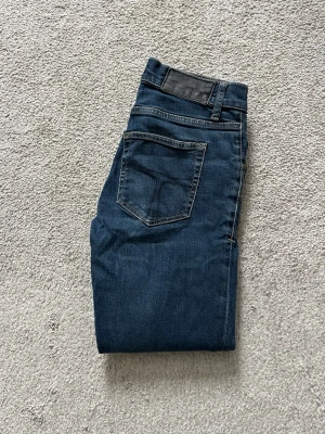 Mörkblå jeans från Tiger of Sweden - Snygga mörkblå jeans från Tiger of Sweden med klassisk femficksdesign och diskret logotyp på linningen. Jeansen har raka ben och är tillverkade i slitstark denim med kontrastsömmar. Perfekta för dig som gillar stilrena och tidlösa plagg.