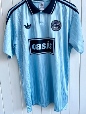 Oasis - Turné 25 - Fotbollströja - Säljer en ljusblå Oasis-supportertröja för bandets turné under 2025. Tröjan är en reprint i nyskick. Aldrig använd. ”25” på ryggen.  Storlek: Large 