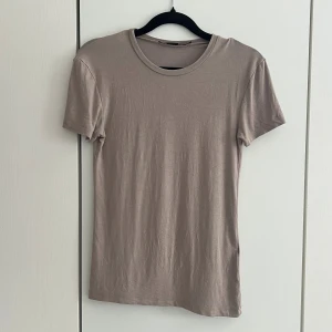Beige basic t-shirt i bomull - En enkel mörkbeige t-shirt med rund hals och korta ärmar. Tillverkad i mjuk bomull och har en normal passform. Perfekt att matcha med jeans eller kjol för en clean och stilren look.