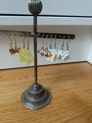 Mixade örhängen med blommor och figurer - Säljer ett set med olika örhängen, bland annat med fjärilar i bronsfärg, ett gult godisformat örhänge med texten 'Sweet', vita blommor, solfjäderform och söta grisar. Materialen varierar mellan metall och plast. Perfekt för dig som gillar unika och lekfulla accessoarer.